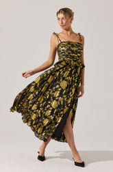 A woman is wearing a sleeveless dress with thin shoulder straps that tie into bows at the shoulders. The dress features a fitted bodice and a high-low skirt with a black and gold floral pattern. The fabric appears to be lightweight and flowing, creating a soft movement.